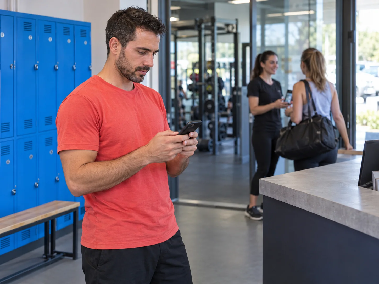 A manager checks his phone at a fitness club while a colleague welcomes a customer.